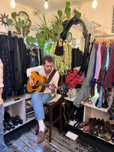 A man "Kipp Boucher" sits at the back of a charity shop on a stool playing an acoustic guitar