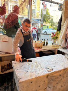 photo of a man in an apron pouring prosecco into glasses on a table covered in a white tablecloth