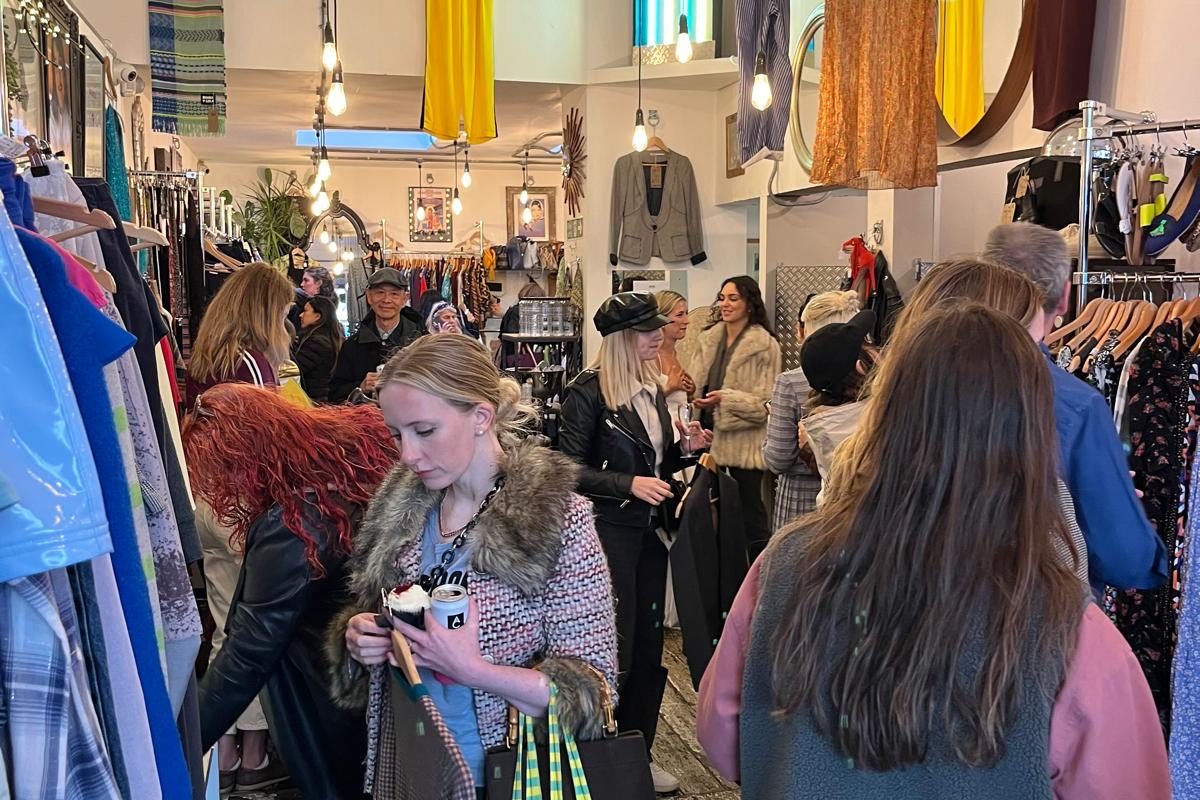 the inside of a charity shop. The walls are lined with rails of clothes and a crowd of people are looking through them and shopping