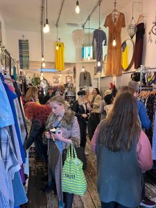 the inside of a charity shop. The walls are lined with rails of clothes and a crowd of people are looking through them and shopping