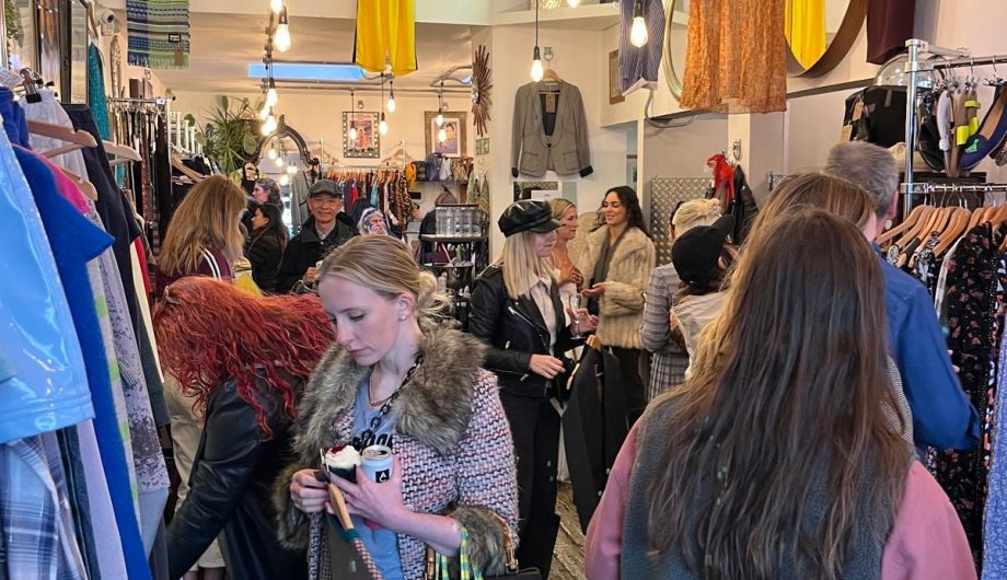 the inside of a charity shop. The walls are lined with rails of clothes and a crowd of people are looking through them and shopping